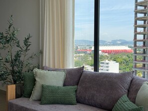 A modern living room with a sofa, a potted plant, and a view of a cityscape through large windows. - Luxury Oceanfront Apartment: Elegance and Relaxation byGPS (Mazatlán)