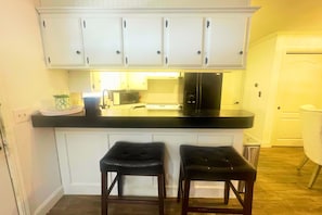 A kitchen with white cabinets, a black countertop, a microwave, a stove, and two black bar stools. - Bourbon & Lace Retreat (Texanna)