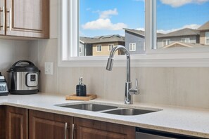 A kitchen with a double sink, a stainless steel kettle, and a window overlooking residential buildings. - Modern 3BR Home | Sleeps 6 | Free Parking & BBQ (Calgary)