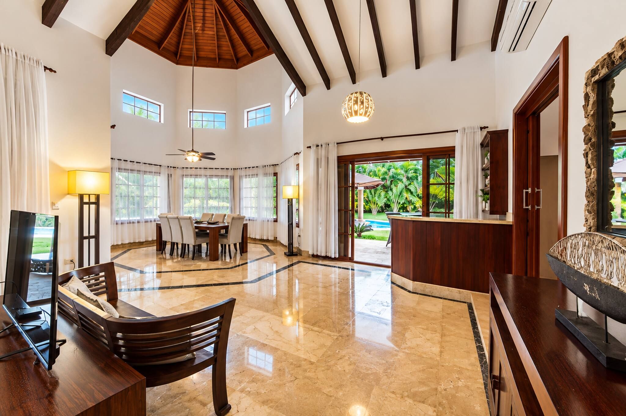 A spacious living area with a high ceiling, a dining table, a TV, and a view of a garden through large windows.