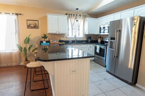 A kitchen with white cabinets, a stainless steel refrigerator, and a central island with stools. - Seahorse House - Coastal Charm, Community Pool & Beach Days (Panama City Beach)