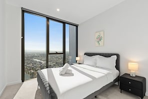 Une chambre moderne avec une grande fenêtre, un lit recouvert de draps blancs, une table de chevet et une lampe fixée au mur.