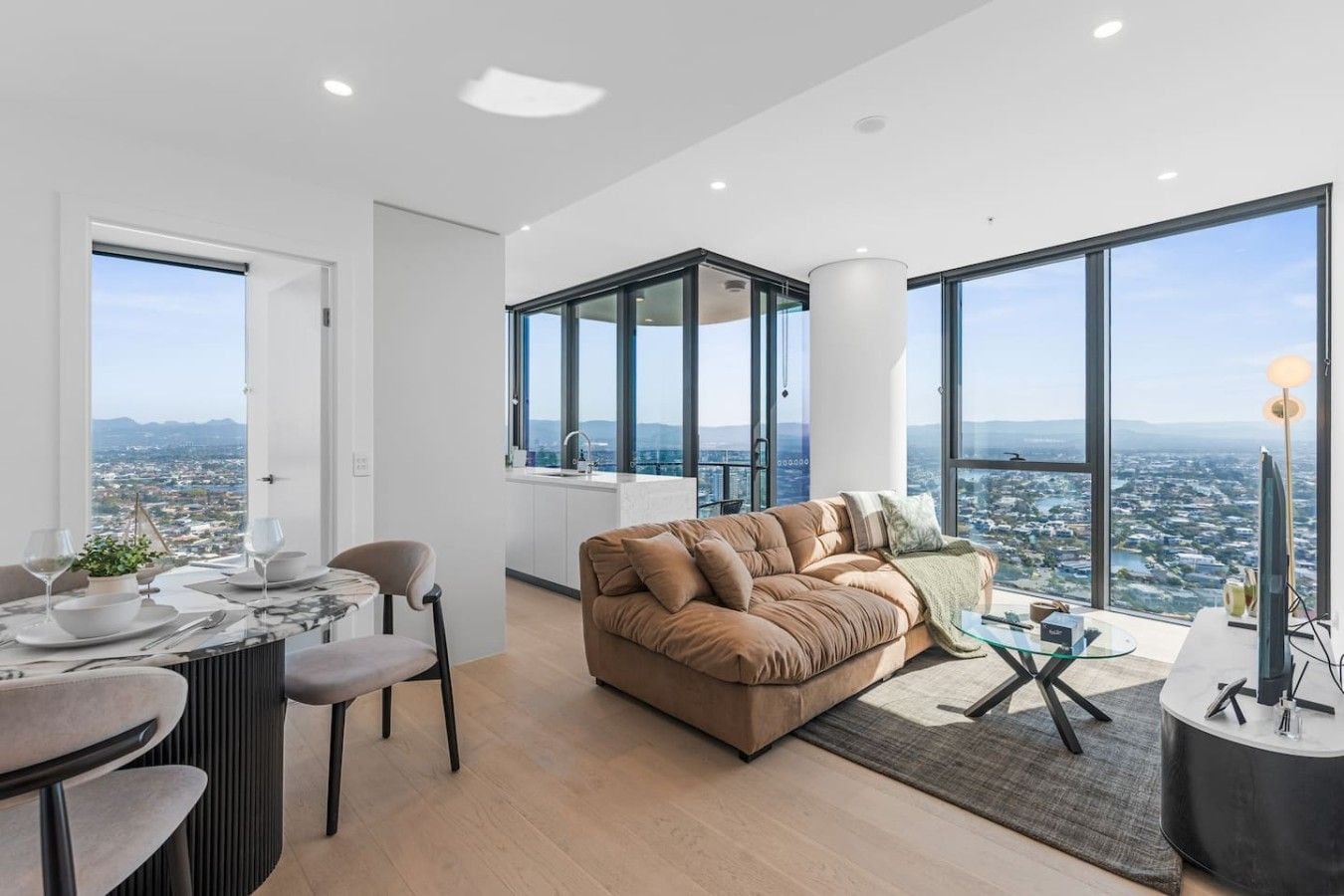 A modern living room with a sofa, dining table, and chairs, large windows with a city view, and a high ceiling.