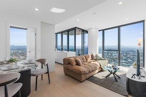 A modern living room with a sofa, dining table, and chairs, large windows with a city view, and a high ceiling. - Luxury 2BR Broadbeach Island Escape (Broadbeach)