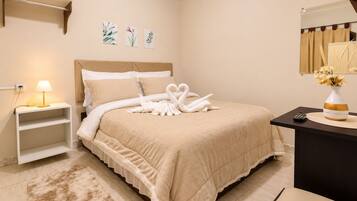 A neatly made bed with a swan towel arrangement, a bedside lamp, a small shelf, and a vase with flowers on a desk.