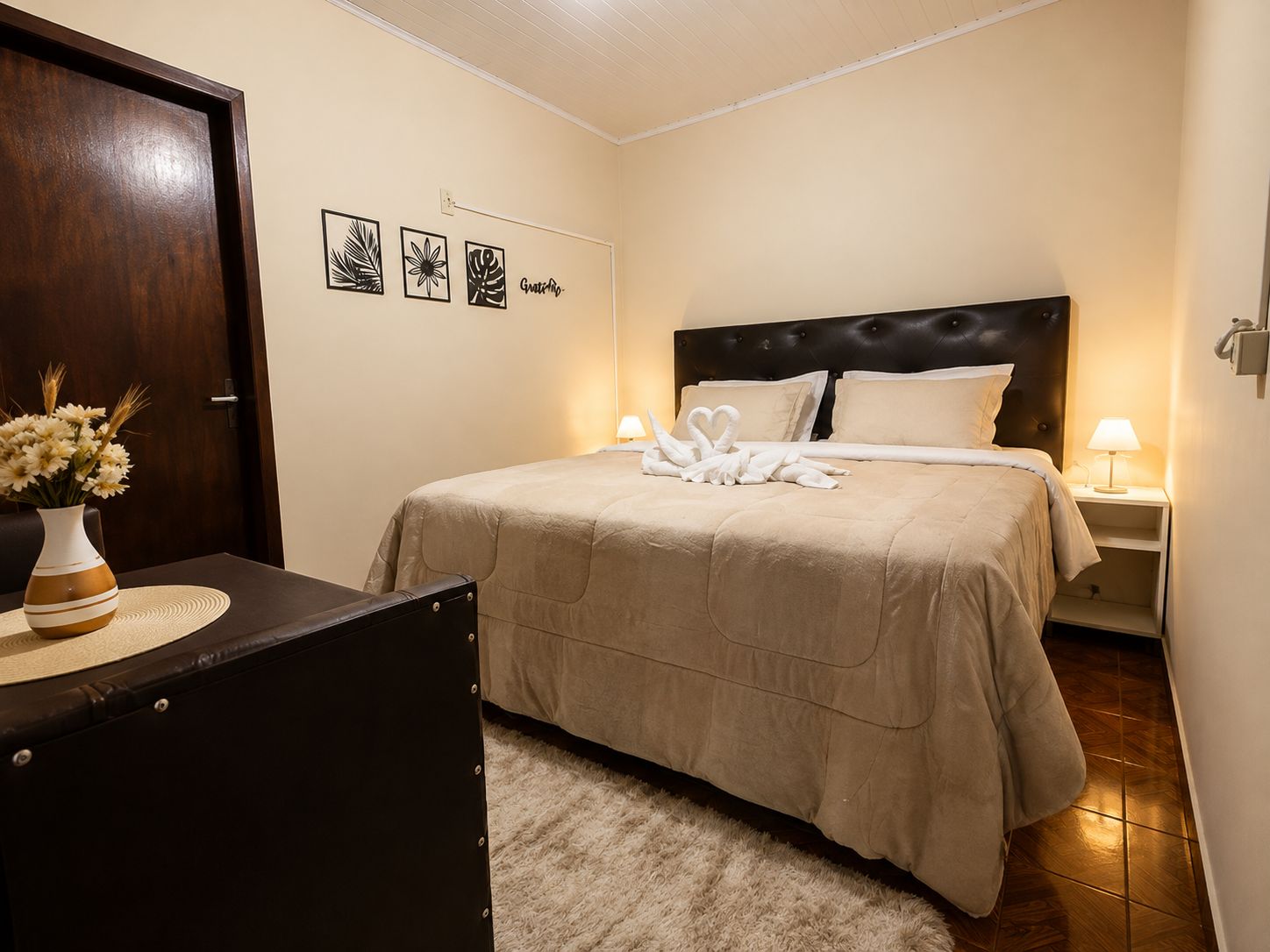 A hotel room with a bed, a nightstand, a lamp, a vase with flowers, and a door.