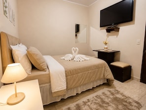 Una habitación de hotel con una cama, una mesita de noche con una lámpara, un televisor montado en la pared y un escritorio pequeño con un jarrón de flores.