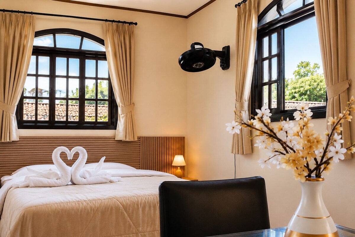 A hotel room with a bed, a table with a vase, and windows with curtains.