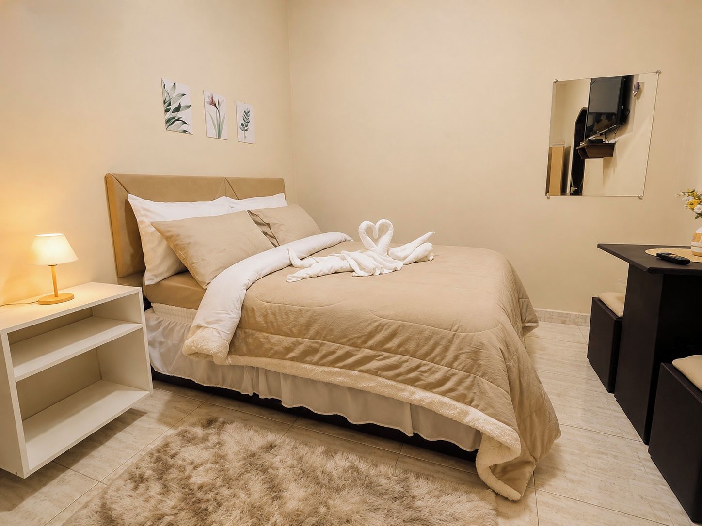 Quarto de hotel com cama, mesa de cabeceira com luminária, tapete macio, escrivaninha com espelho e obras de arte na parede.