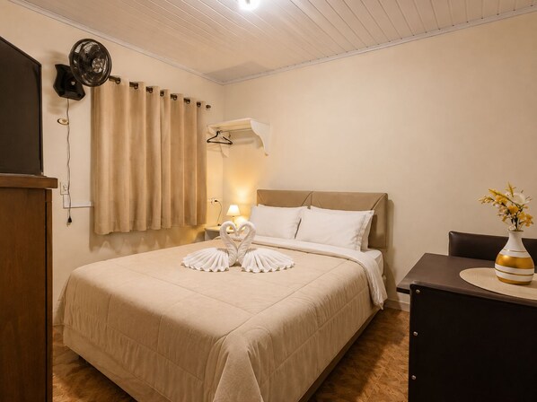 A hotel room with a bed, a bedside table, a lamp, a wall-mounted fan, and a window with curtains.