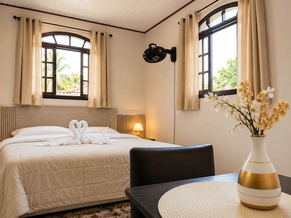 Habitación de hotel con cama, ventilador de pared, mesa con un florero y dos ventanas con cortinas.
