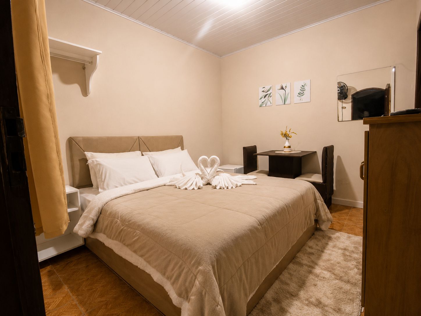 A hotel room with a bed, a bedside table with a vase of flowers, a small table with a fan, and a mirror on the wall.