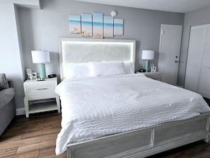 A neatly arranged bedroom with a large bed, two bedside tables, a nightstand, a lamp, and a wall-mounted picture. - 3 Oceanview Studios connecting w/pool, tiki bar (Daytona Beach)