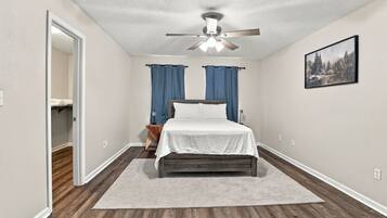 A bedroom with a bed, a ceiling fan, a painting, and a view into another room.