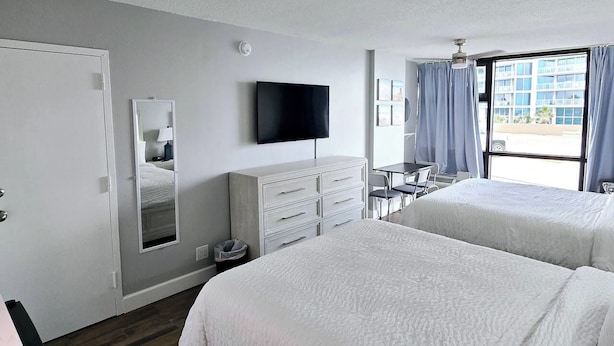 A hotel room with two beds, a flat-screen TV, a desk, and a window with curtains. - New updated Daytona Oceanview w/kitchenette, pool (Daytona Beach)