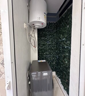 A compact bathroom with a water heater mounted on the wall, a small storage unit, and a wall covered with artificial green foliage. - 5 min to Mohammed VI Hospital + 15 min to the airport (Bouskoura)