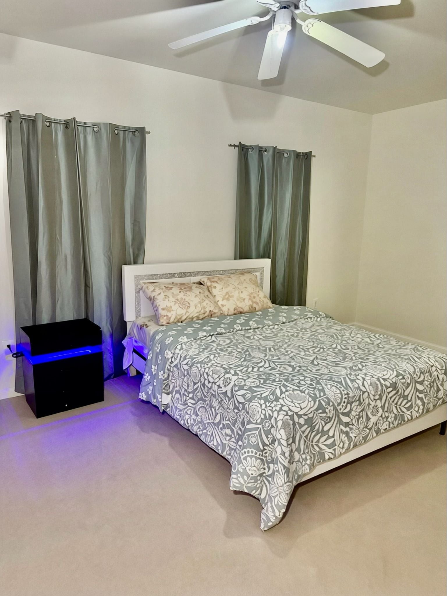A bedroom with a bed, curtains, a ceiling fan, and a small black object on the floor.