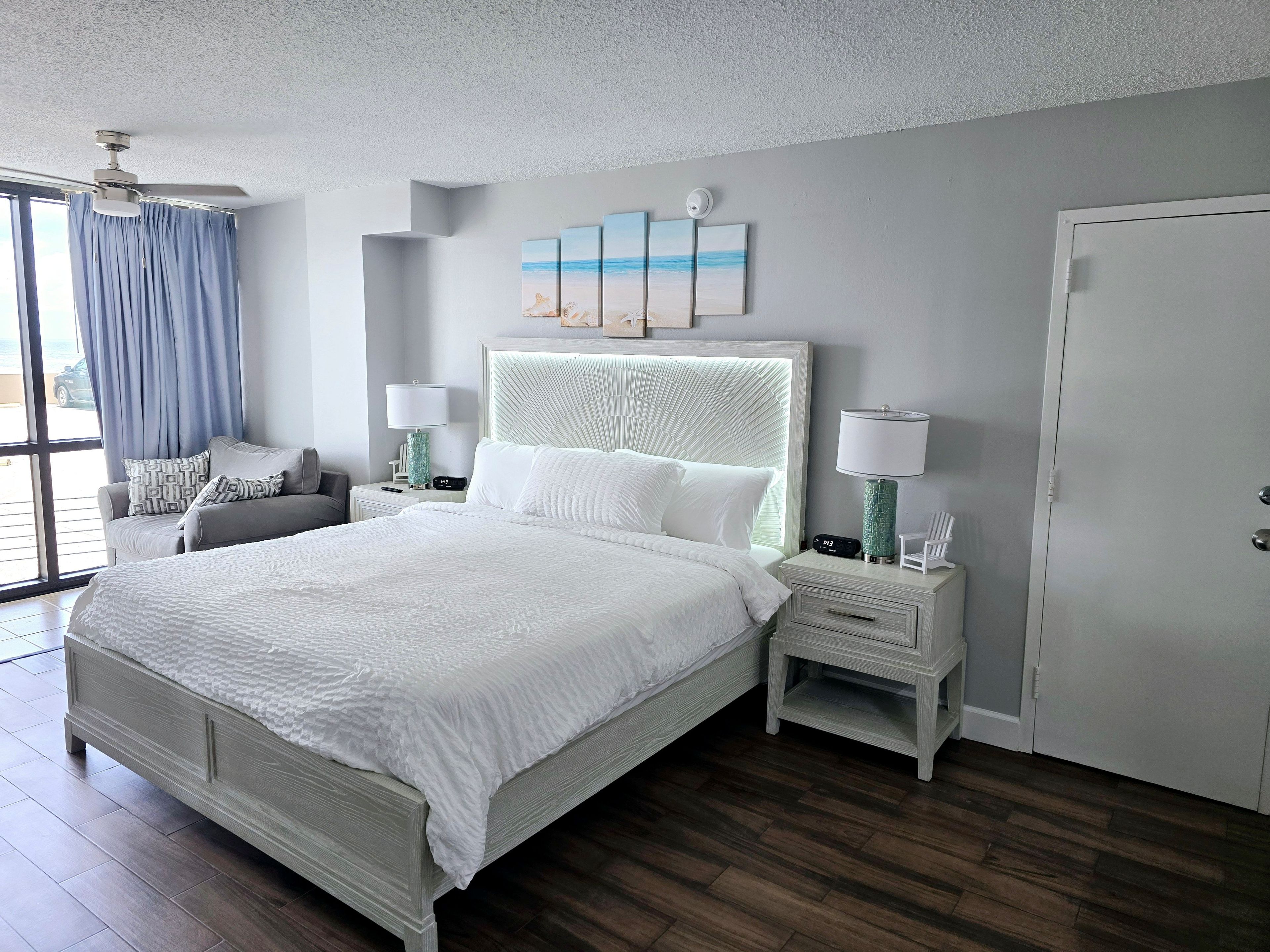 Un dormitorio con una cama grande, una mesita de noche con lámpara, una silla, una ventana con cortinas y una puerta.