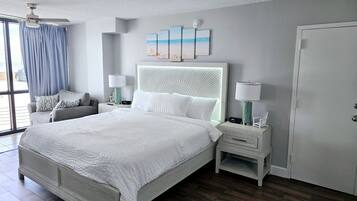 Un dormitorio con una cama grande, una mesita de noche con lámpara, una silla, una ventana con cortinas y una puerta.