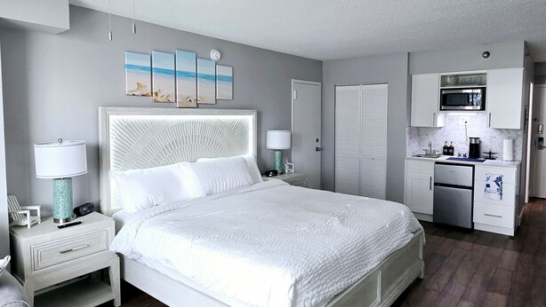 Une chambre à coucher comprenant un lit, des tables de chevet, un ventilateur de plafond, des œuvres d’art, un placard et un coin cuisine.