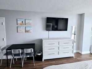 A room with a flat-screen TV mounted on the wall, a white dresser with drawers, two chairs, and a small trash bin. - Spectacular New King Studio w/kitchenette, pool (Daytona Beach)