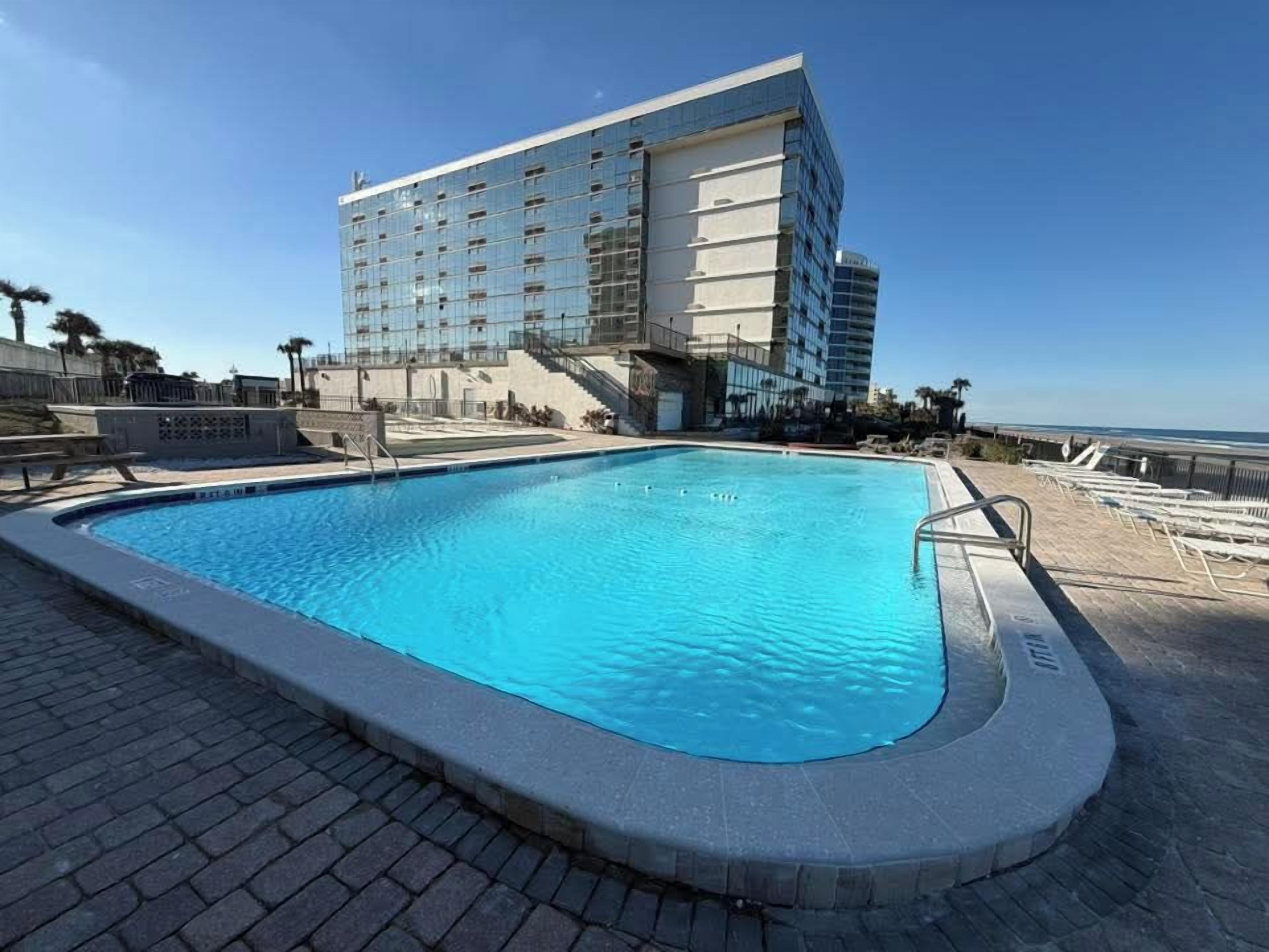 A swimming pool with a clear blue water surface, surrounded by a paved deck. In the background, there is a large hotel building with multiple floors and a glass facade.