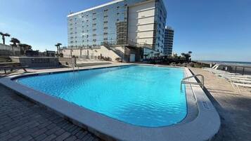 A swimming pool with a clear blue water surface, surrounded by a paved deck. In the background, there is a large hotel building with multiple floors and a glass facade.