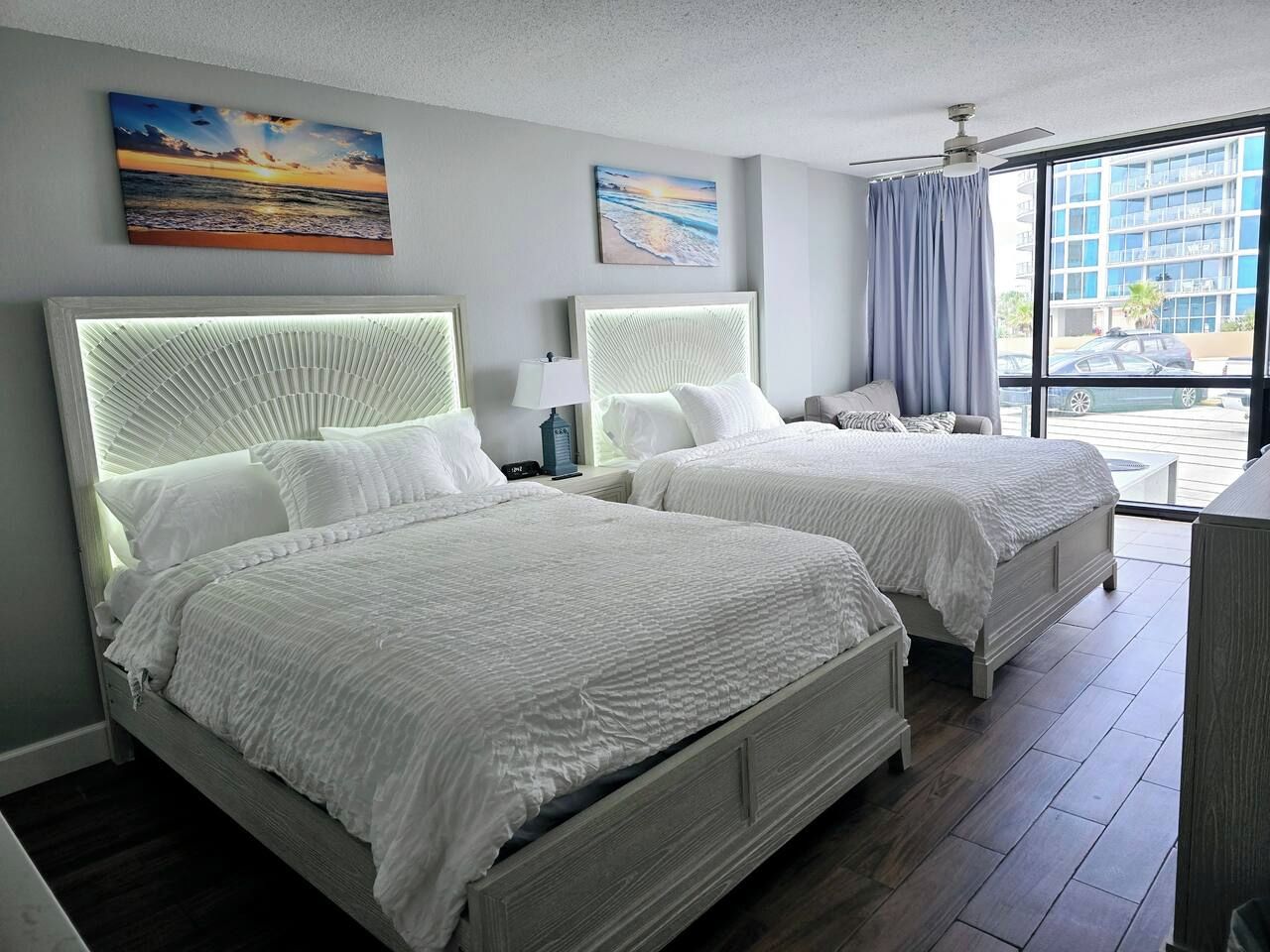 Habitación de hotel con dos camas, ventilador de techo y ventanas grandes con vista a la ciudad.