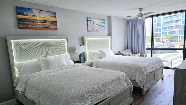 Quarto de hotel com duas camas, ventoinha no teto e amplas janelas com vista para a cidade.