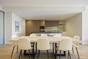 A modern kitchen with a central island, a dining area with a large table and chairs, and a framed picture on the wall. - Starboard Cove (Safety Beach)