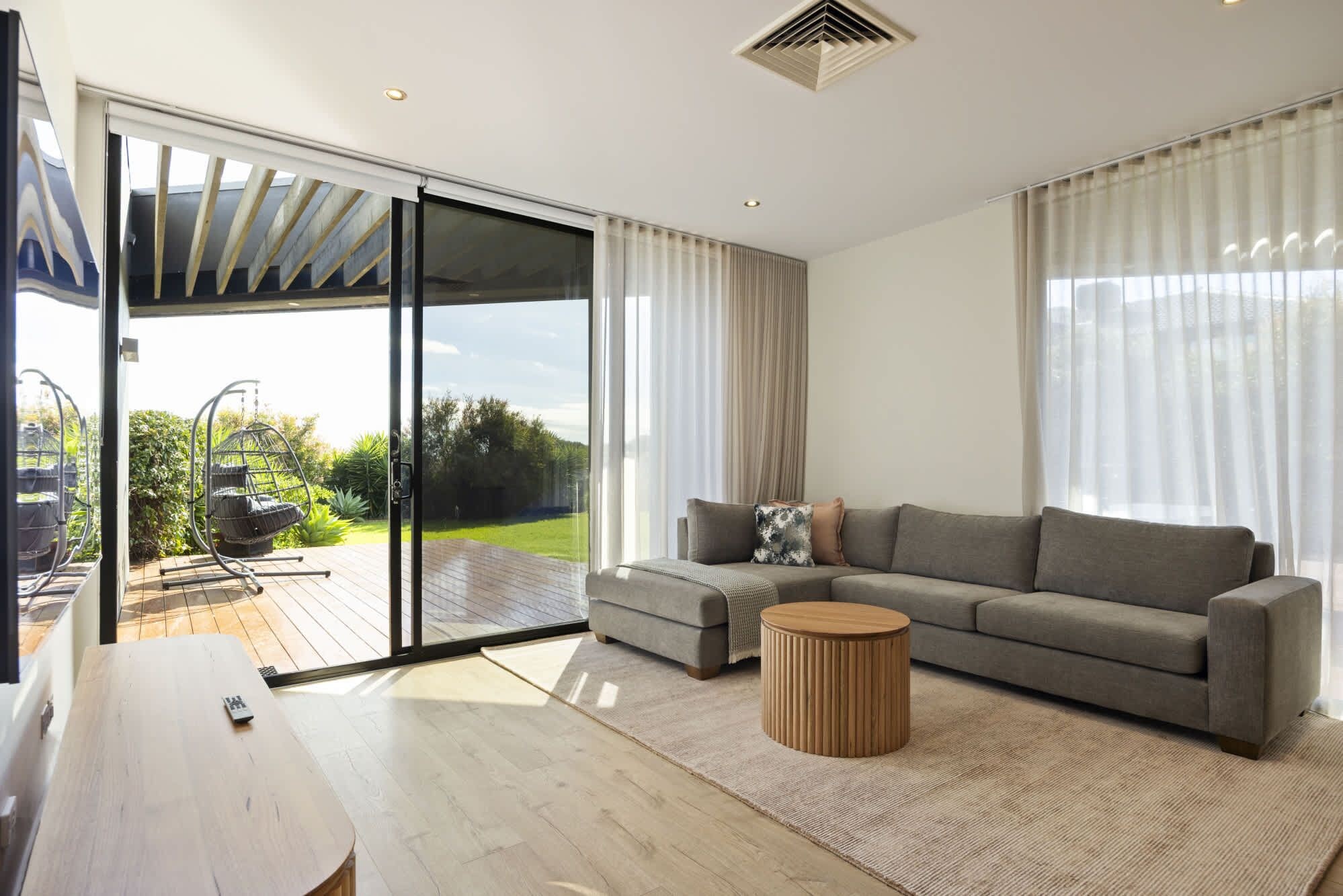 Ein modernes Wohnzimmer mit einer Schiebeglasdoor zum Patio, einem grauen Sofa, einem runden Couchtisch aus Holz und einem Holzboden.