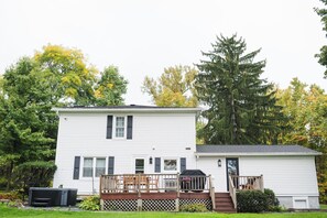 A two-story white house with a wooden deck and a covered porch. - Garden Gate Farmhouse- a peaceful retreat with hot tub and hammocks! (Skaneateles)