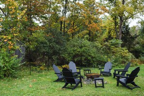 Eine Gruppe Adirondack-Stühle ist um eine Feuerstelle in einem grünen Außenbereich mit Bäumen im Hintergrund angeordnet.