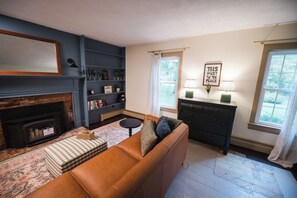 A living room with a fireplace, a sofa, a bookshelf, and a window with curtains. - Garden Gate Farmhouse- a peaceful retreat with hot tub and hammocks! (Skaneateles)
