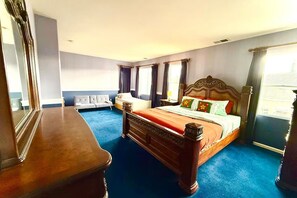 A hotel room with a large bed, a wooden dresser, a white armchair, and a blue carpet.