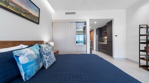 A bedroom with a large bed, a nightstand, a bedside table, a bookshelf, and a view of the kitchen and balcony. - One 503B Studio: Vista del Sol Studio (Puerto Peñasco)