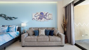 Een moderne slaapkamer met een grijze bank, een bed met blauw beddengoed, een kunstwerk aan de muur en uitzicht door een groot raam.