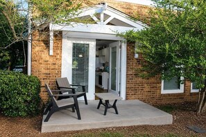 Une maison en briques avec un patio couvert, un coin repas avec des chaises et une cuisine visible à travers une porte ouverte.