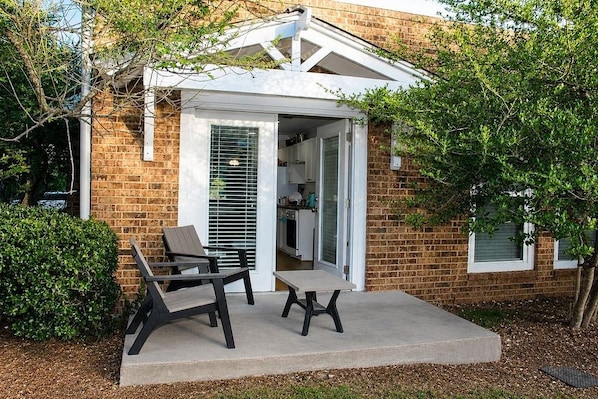 A brick house with a covered patio, a dining area with chairs, and a kitchen visible through an open door.