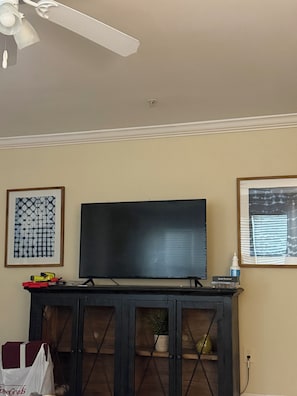 A living room with a flat-screen TV, a ceiling fan, and framed artwork on the walls.