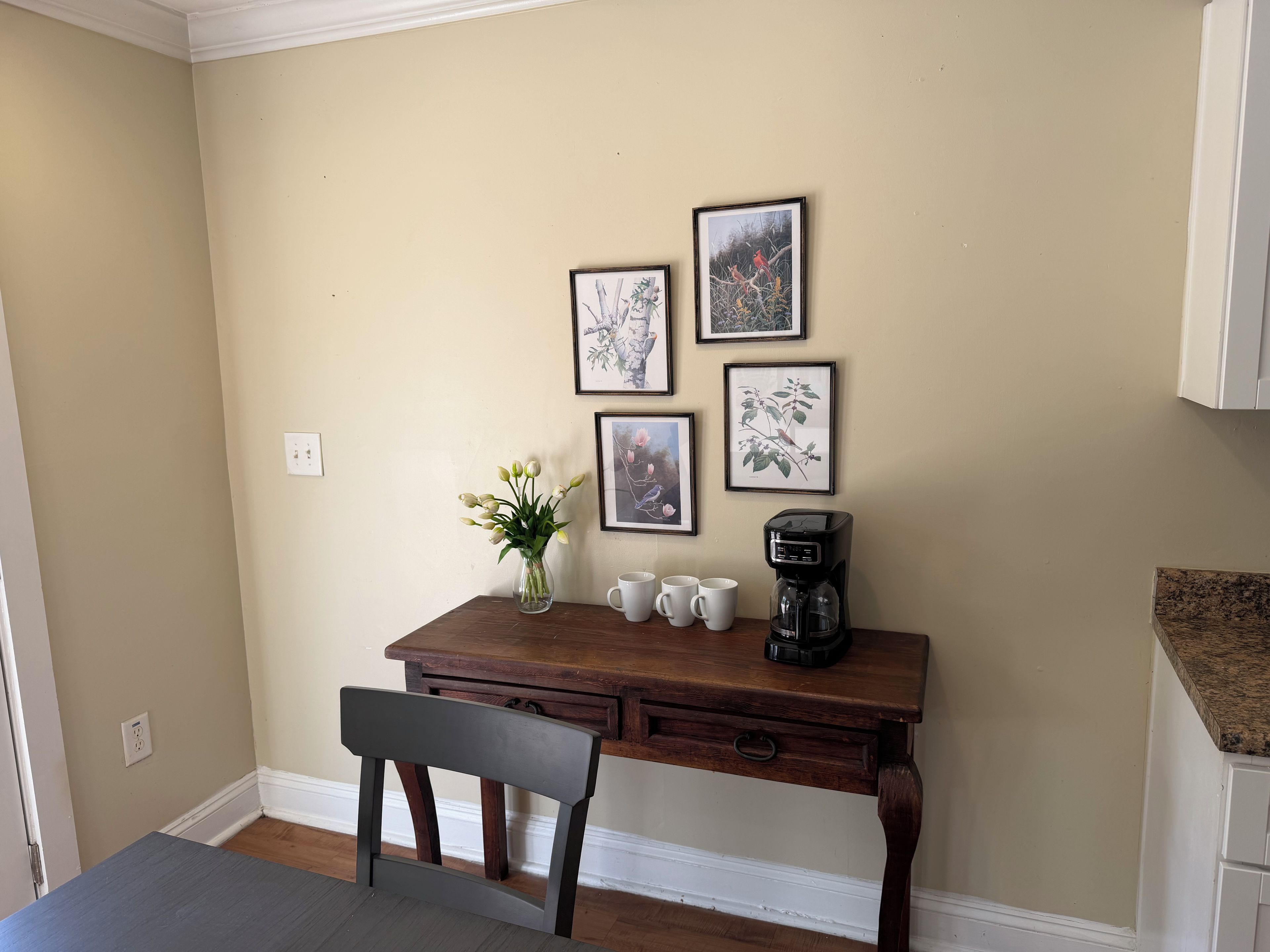 Un petit coin repas avec une table en bois, un vase de fleurs, une machine à café et, au mur, des reproductions encadrées de gravures botaniques.