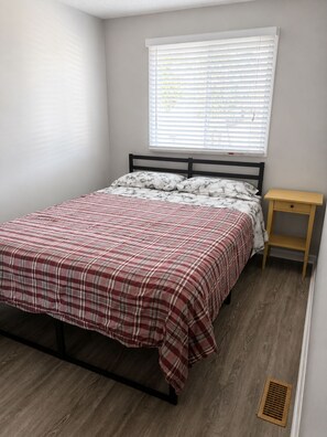 Ein Schlafzimmer mit Bett, einem Fenster mit Jalousien, einem Nachttisch und Holzboden.