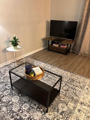 Ein Wohnzimmer mit einem schwarzen Couchtisch, einem weißen Beistelltisch mit Pflanze, einem TV-Schrank mit Flachbildfernseher und einem gemusterten Teppich.
