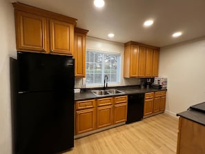 Una cocina con armarios de madera, un refrigerador negro, un lavamanos de acero inoxidable y una ventana con persianas.