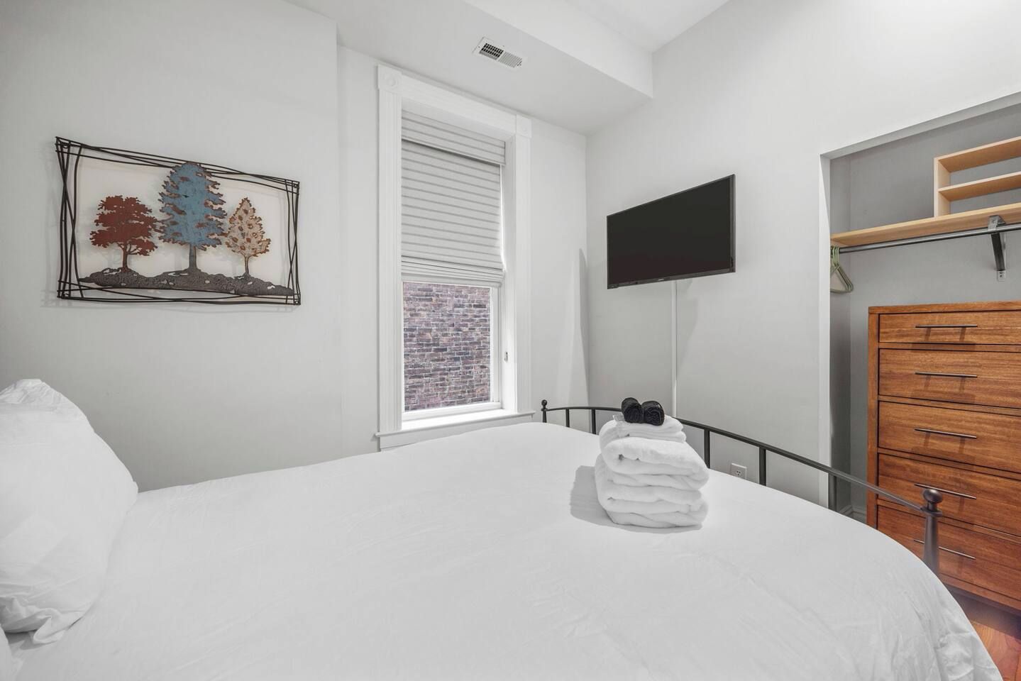 A bedroom with a bed, a TV mounted on the wall, a window with blinds, and a wooden dresser.