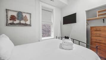 A bedroom with a bed, a TV mounted on the wall, a window with blinds, and a wooden dresser.