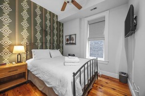 Un dormitorio con cama, mesita de noche, lámpara, ventana y televisor montado en la pared.