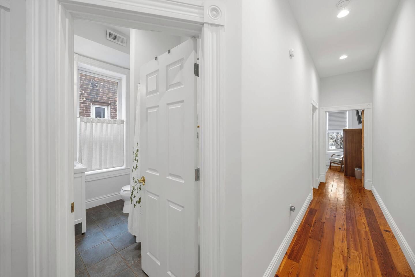 A hallway with a wooden floor, white walls, and a white door with a window.