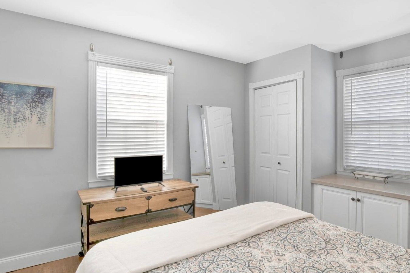 A bedroom with a bed, a dresser, a mirror, and two windows with blinds.