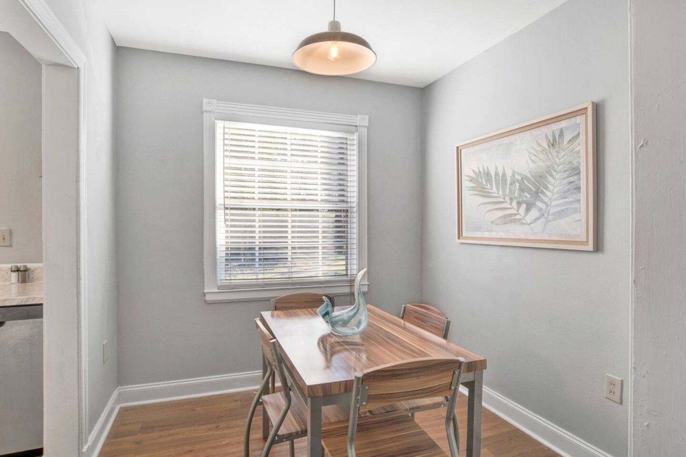A dining area with a wooden table and chairs, a window with blinds, and a framed artwork on the wall.